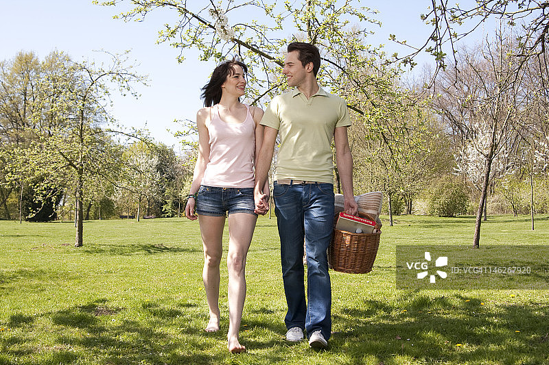 年轻夫妇手牵手挎着野餐篮在草地上散步图片素材