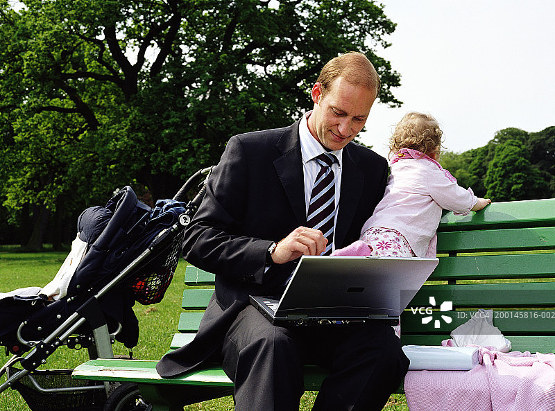 商人和女儿(12-15个月)坐在公园长椅上，使用笔记本电脑图片素材