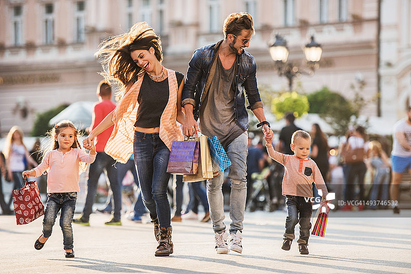 年轻快乐的家庭带着购物袋在街上奔跑。图片素材