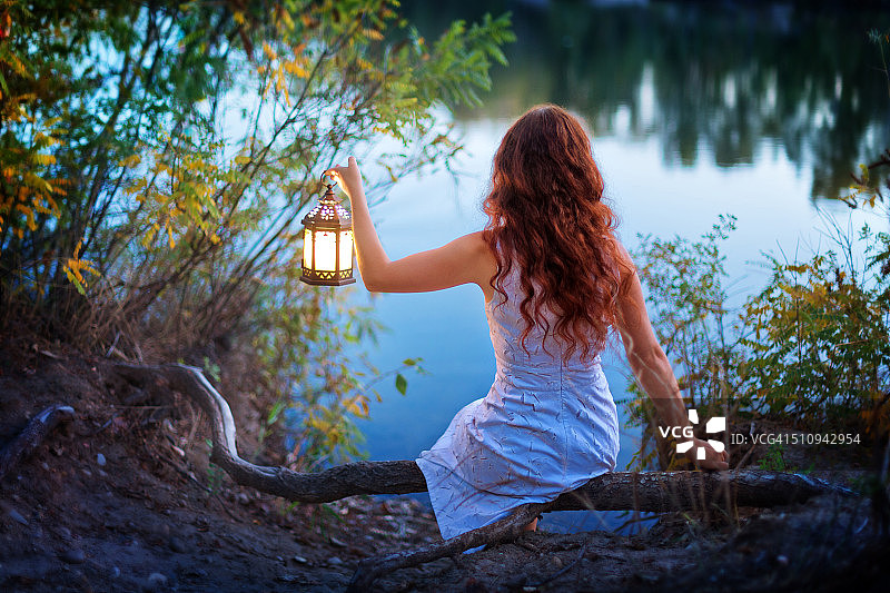 黄昏时分，坐在湖边，身着白色无袖连衣裙，挽着小灯笼，一头红卷发的年轻女子的背影图片素材