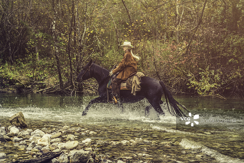 女牛仔骑着马穿过森林里的一条河图片素材