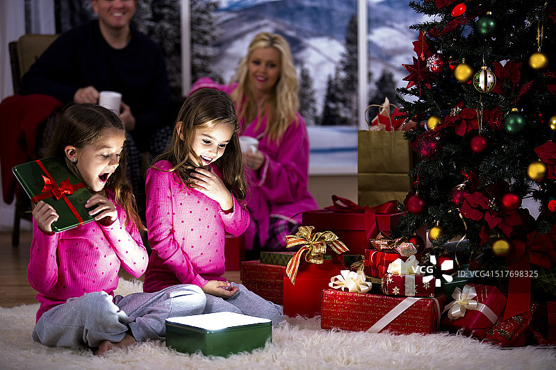 女孩们兴奋地打开圣诞礼物。父母，树，礼物背景。图片素材
