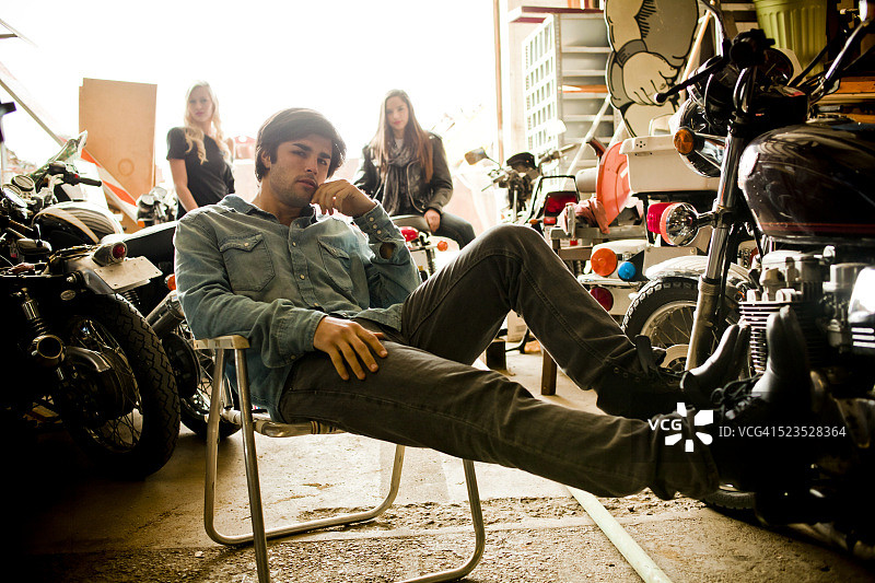 一名男子坐在摩托车车库的折叠椅上休息，后面站着两名女子图片素材