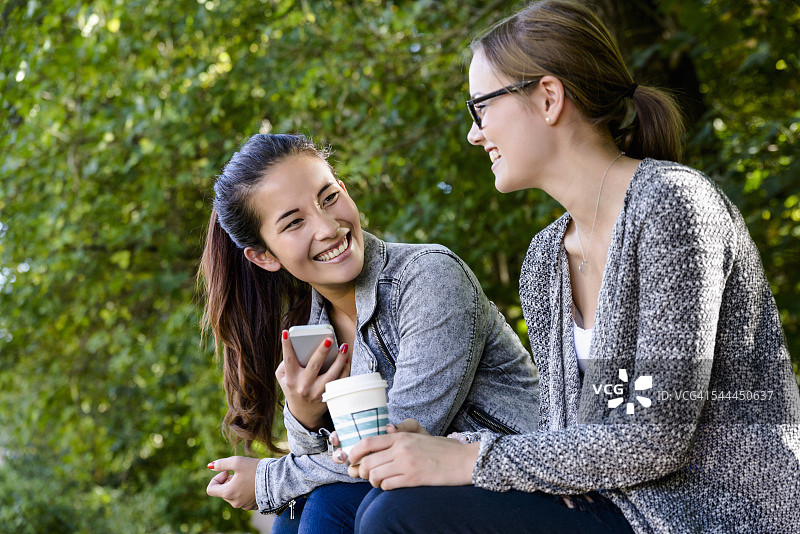 两个年轻女子端着外卖咖啡坐在公园里聊天图片素材