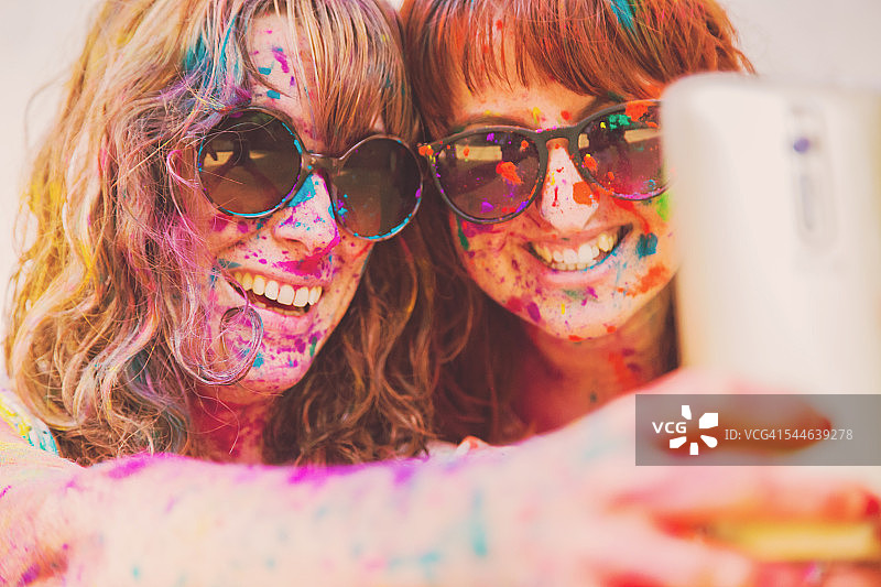 在一场胡里节派对上，两个年轻女孩用涂有彩色Gulal粉末的智能手机微笑着自拍，以欢迎周日家庭聚会上的春天。图片素材