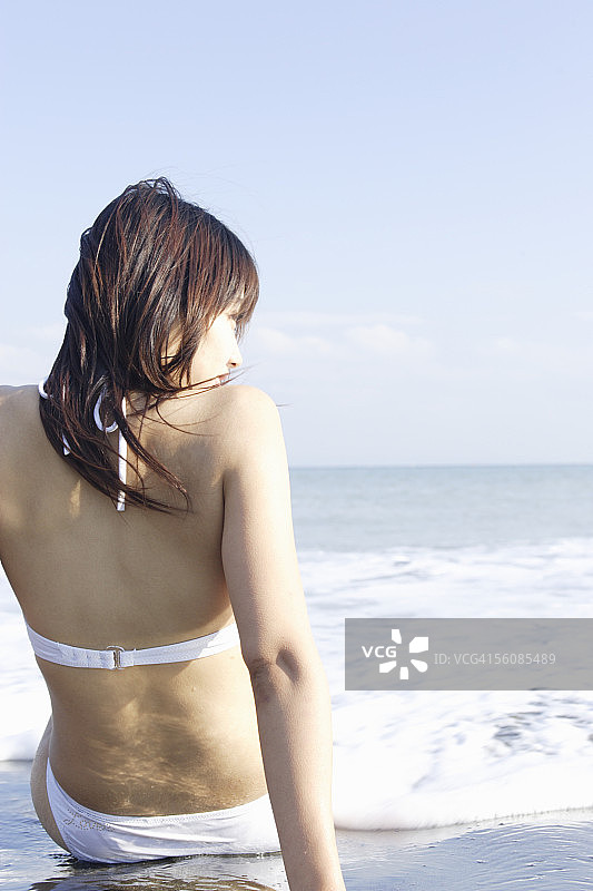 一个穿着比基尼的年轻女子坐在沙滩上，后视图图片素材