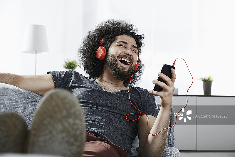 大笑的年轻人躺在家里的沙发上，用耳机和智能手机听音乐图片素材