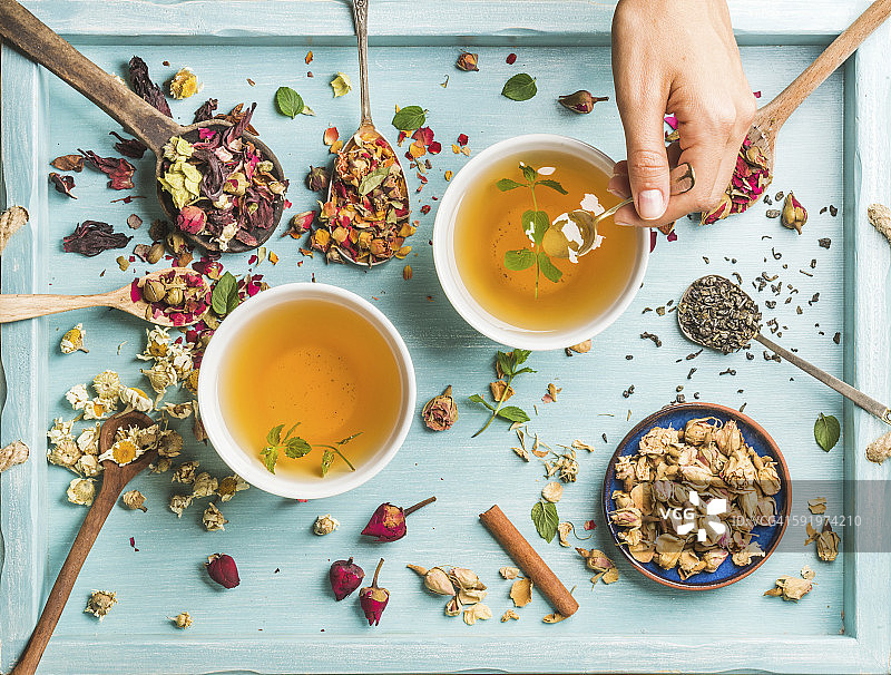 两杯健康的草药茶与薄荷，肉桂，干玫瑰和甘菊花在勺子和男人的手握匙蜂蜜图片素材