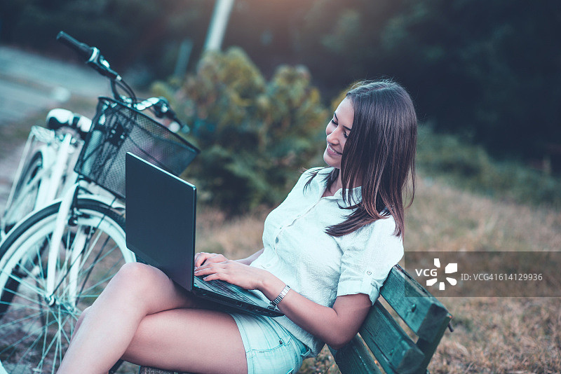 微笑的女人坐在长椅上，骑完自行车后使用笔记本电脑图片素材