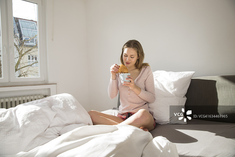 年轻女子坐在床上，端着咖啡和羊角面包图片素材
