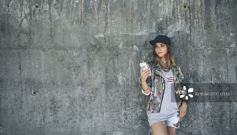 年轻女子靠在水泥墙上，戴着耳机听音乐图片素材