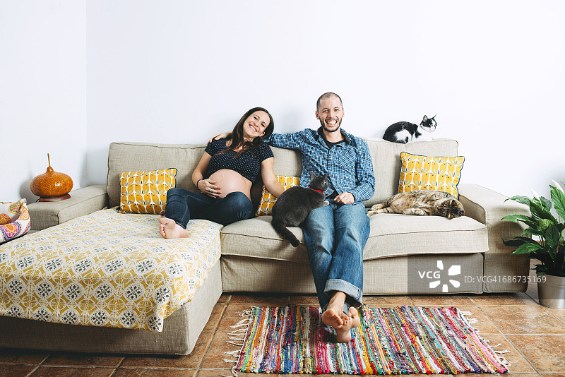 一对幸福的夫妇期待着一个宝宝和三只猫坐在家里的沙发上图片素材