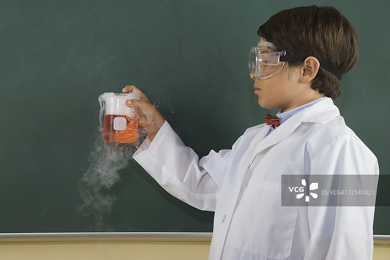 男孩穿着实验服，戴着护目镜，拿着热气腾腾的烧杯在教室里图片素材