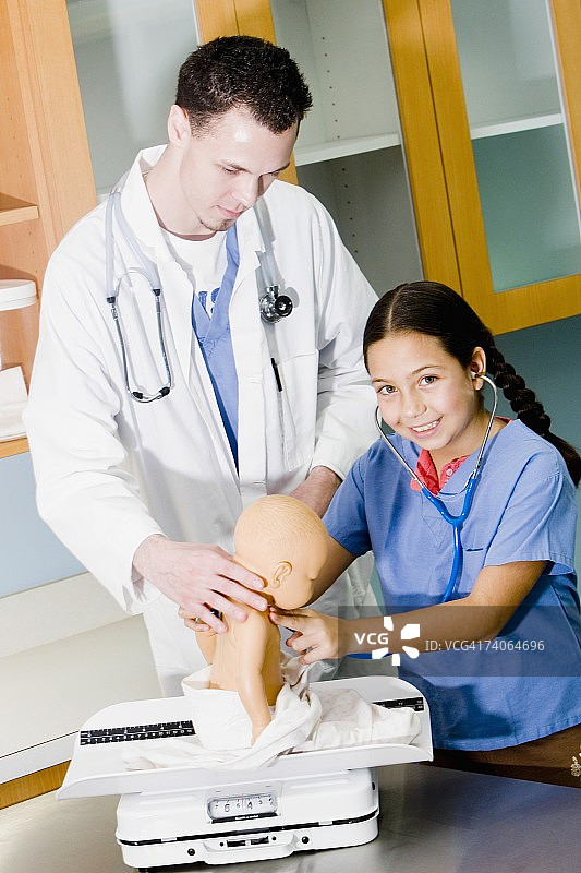 一个女孩正在用听诊器检查一个洋娃娃，旁边站着一位男医生图片素材