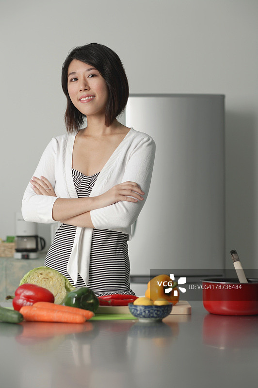 年轻女人在厨房里，双臂交叉，蔬菜放在她面前的桌子上图片素材