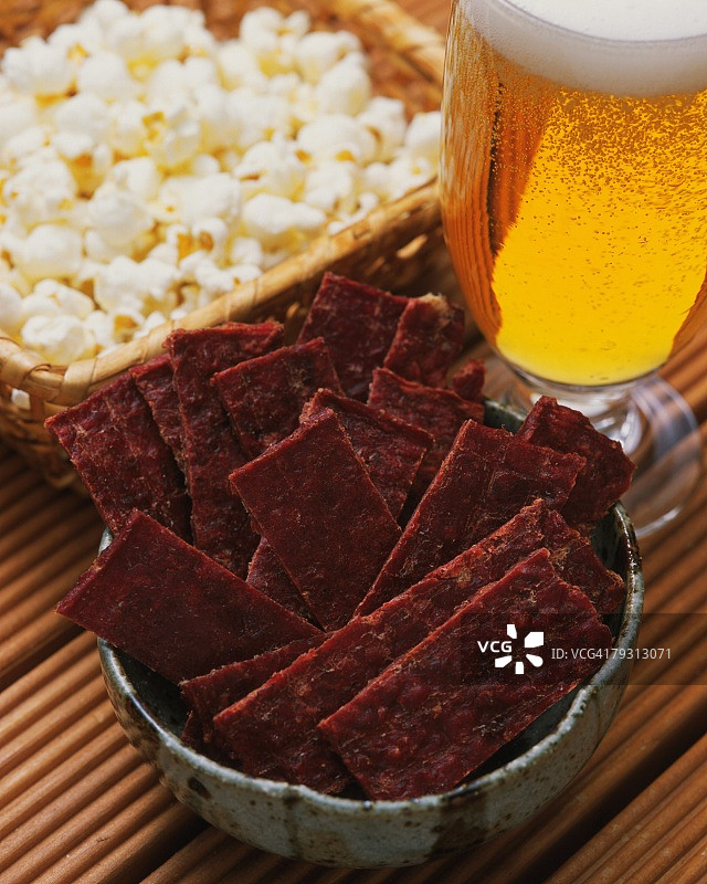 一个小碗装满牛肉干，和一个装满爆米花的木篮子旁边的一杯啤酒，高角度的观点图片素材