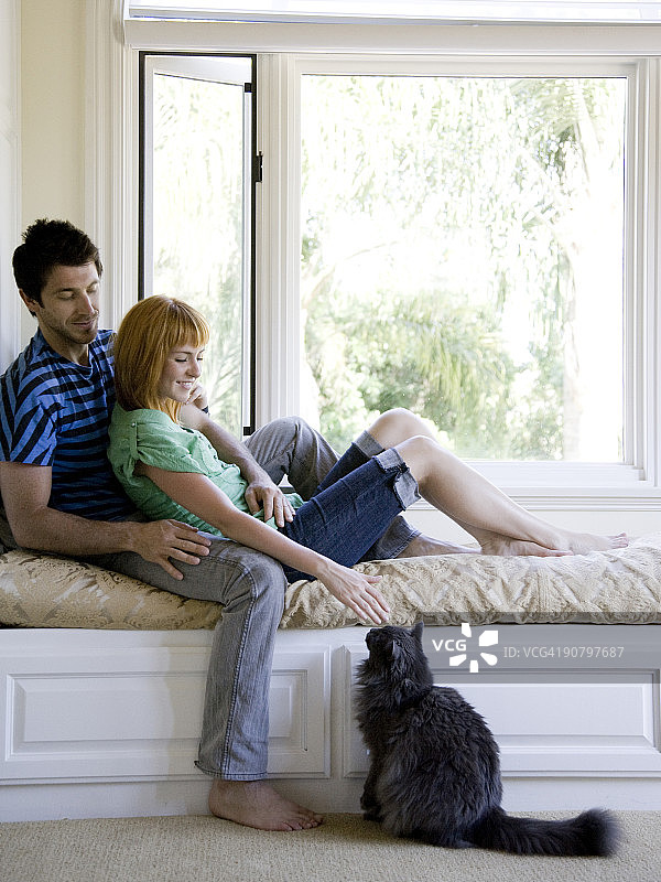 一对夫妇坐在靠窗的长椅上抚摸着一只猫图片素材