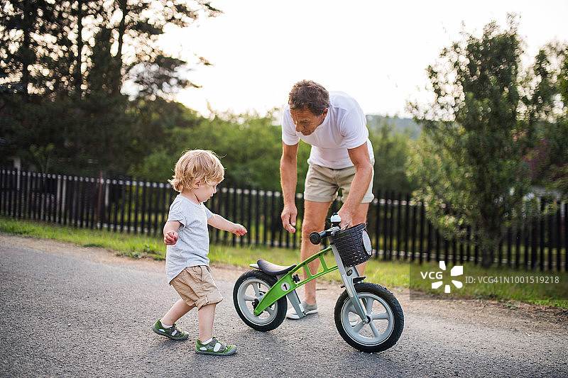 一个蹒跚学步的小男孩和他的祖父在户外抱着一辆儿童平衡自行车。图片素材