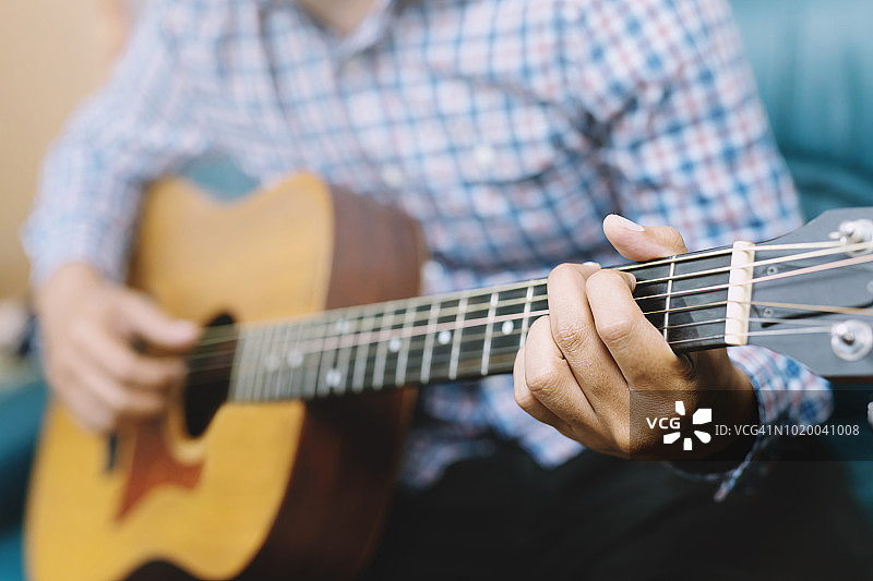 时髦的家伙手弹原声吉他。艺术家年轻的男音乐家格子衬衫演奏吉他在沙发客厅客厅。图片素材