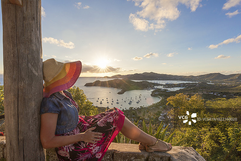 在安提瓜岛的雪莉高地上，一位戴着迷人的帽子和衣服的女士在欣赏英国港口上浪漫的日落图片素材