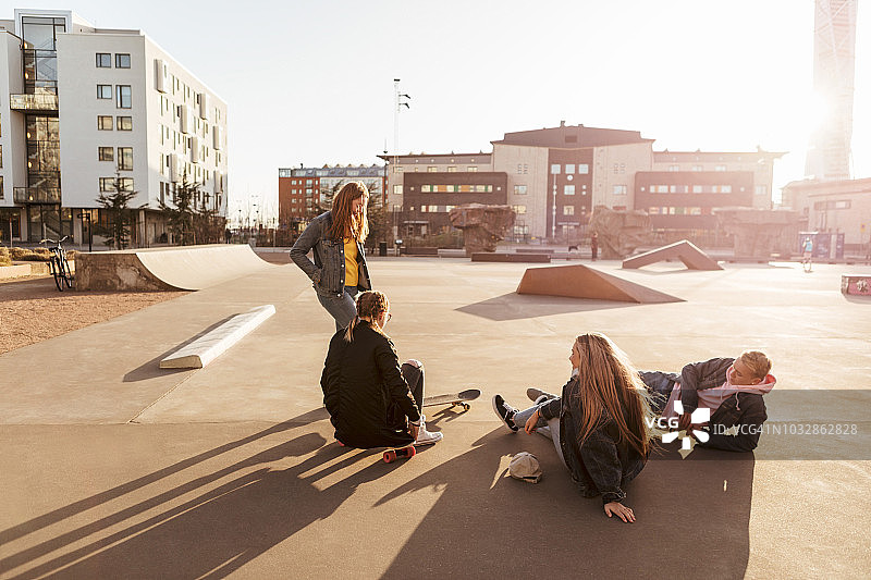 在阳光明媚的日子里，女孩站着和坐在滑板公园的青少年朋友聊天图片素材