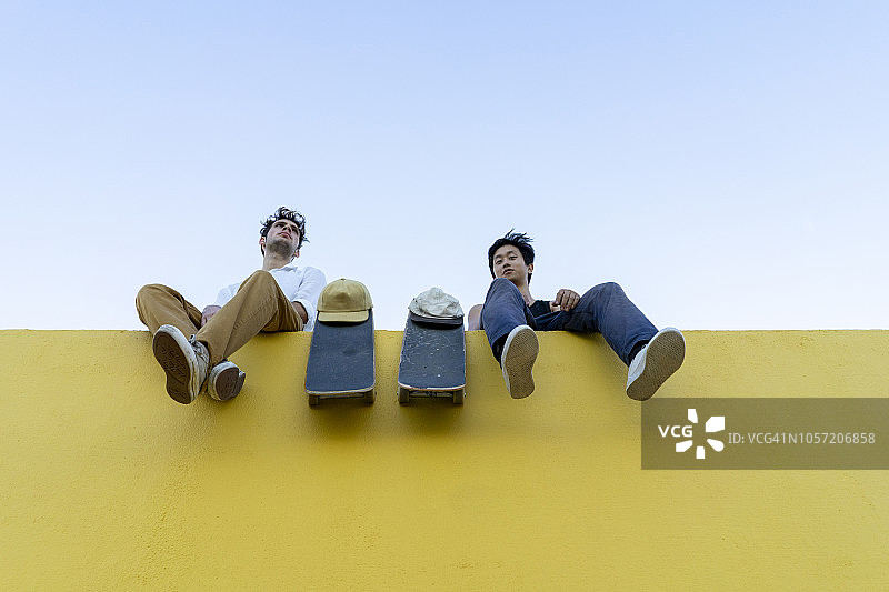 两个拿着滑板的年轻人坐在一堵黄色的高墙上图片素材