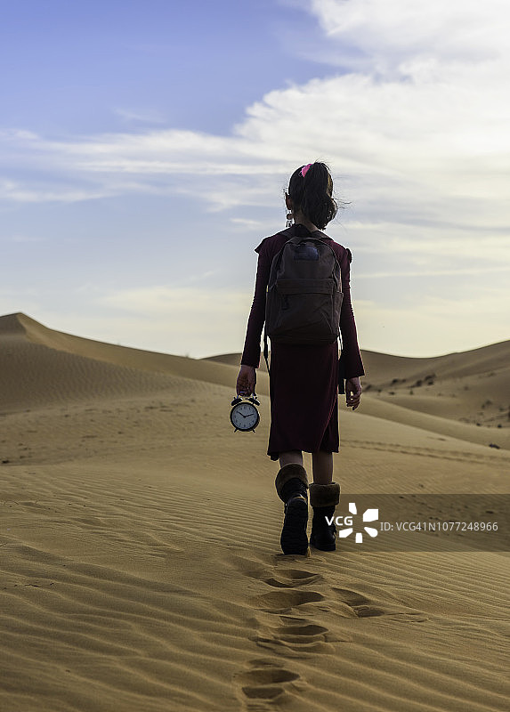 一个孤独的女孩在沙漠中走着手里拿着一个闹钟。图片素材
