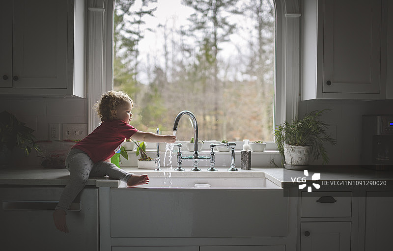 可爱的女孩坐在厨房柜台上靠窗的时候在水槽里洗手的侧视图图片素材