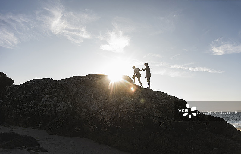 法国，布列塔尼，一对年轻夫妇在日落时分在海滩上爬岩石图片素材