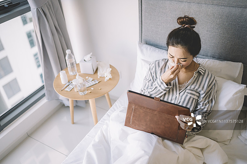 一位亚洲华裔女性在床上穿着睡衣，用纸巾捂着鼻子看数字平板电脑图片素材