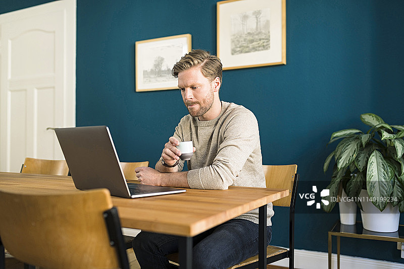 一个男人在家庭办公室的木桌上用笔记本电脑工作，喝着浓咖啡图片素材