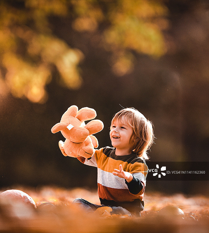 快乐的小男孩在秋天的树叶里玩泰迪熊。图片素材