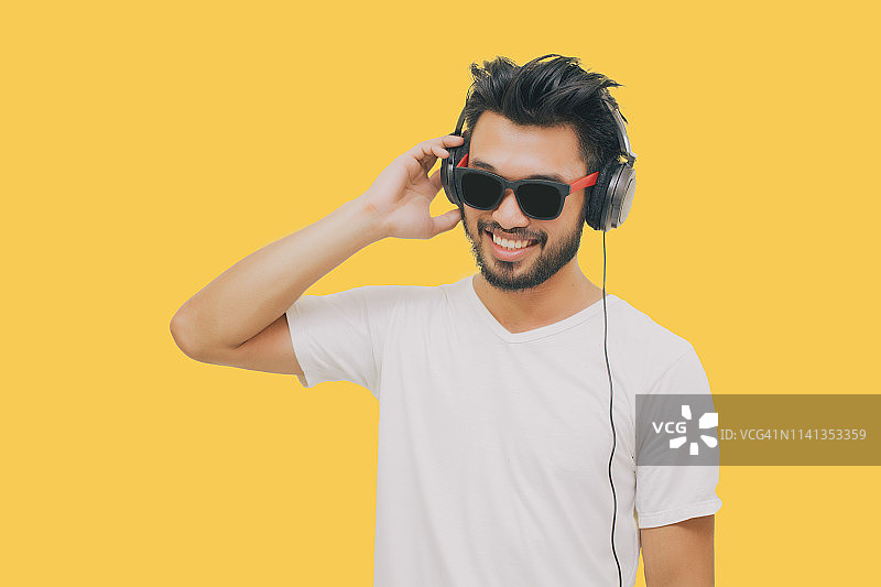 亚洲英俊的男人，有胡子，微笑和大笑，用智能手机听音乐的黄色背景耳机图片素材