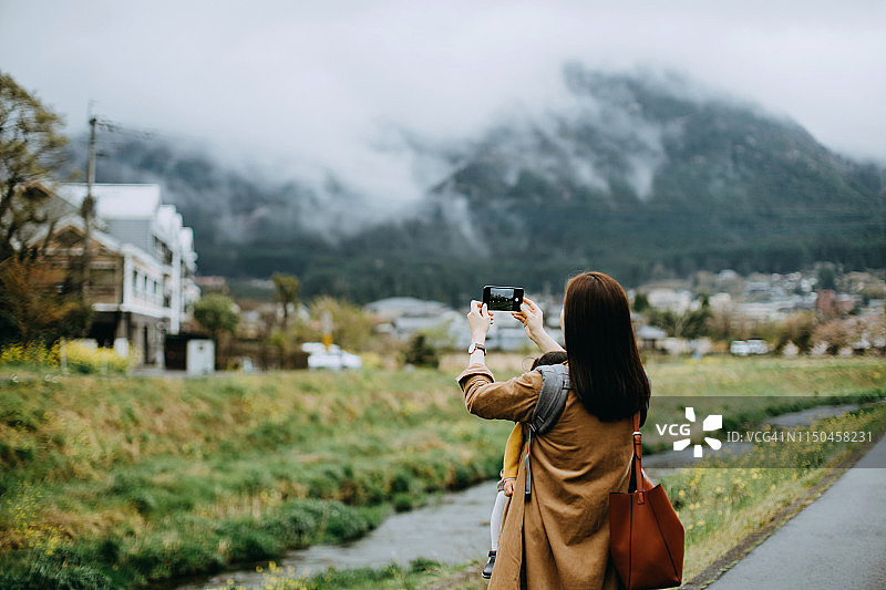 年轻的亚洲母亲抱着蹒跚学步的小女孩在户外散步，度假时用智能手机拍摄美丽的自然风景图片素材