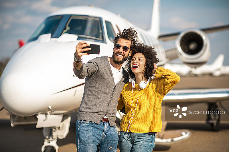 一对富有的年轻夫妇正在机场停机坪上的一架私人飞机前用手机自拍图片素材