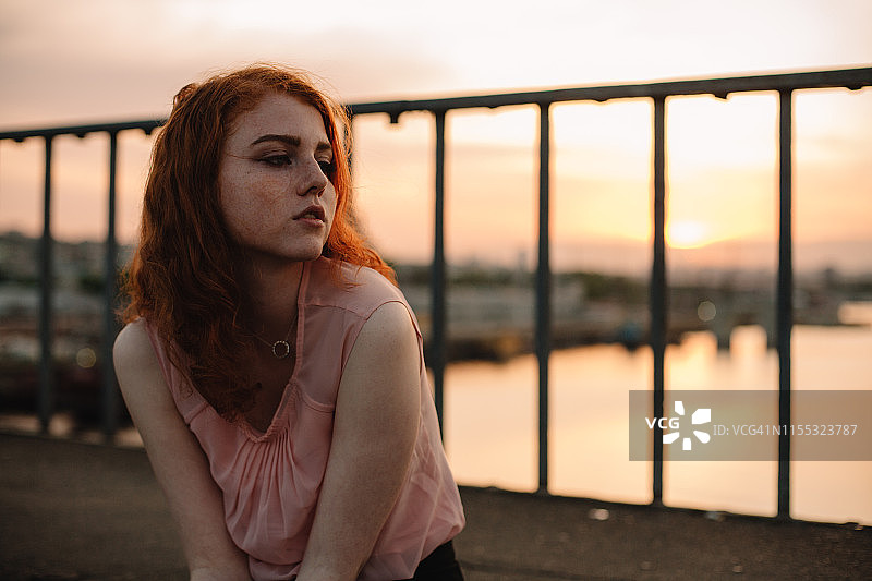 日落时分，年轻的红发女子坐在桥上的肖像图片素材