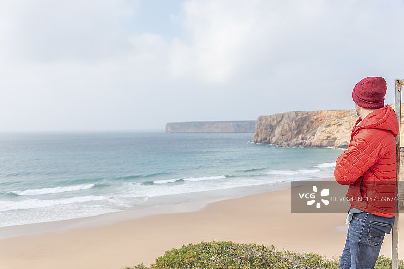 葡萄牙，阿尔加维，萨格雷斯，普拉亚·多·贝利切，一名男子戴着红色的帽子，穿着红色的夹克，看着海滩和大海图片素材