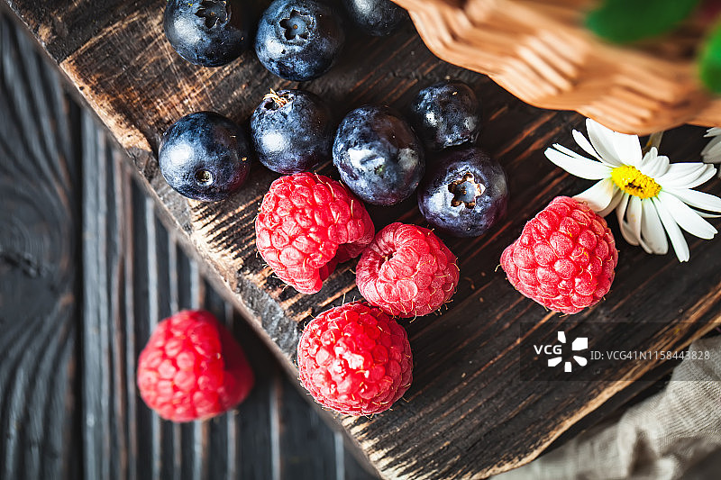 覆盆子和蓝莓装在一个篮子里，洋甘菊和叶子在黑暗的背景上。夏日与健康的饮食理念。有选择性的重点。平的。图片素材