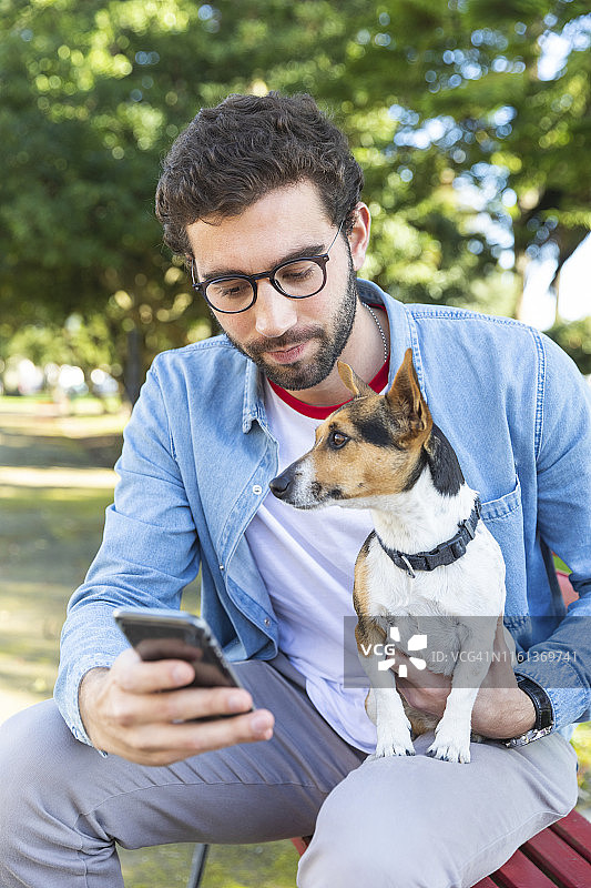 一个年轻人和他的狗坐在公园长椅上看智能手机的照片图片素材