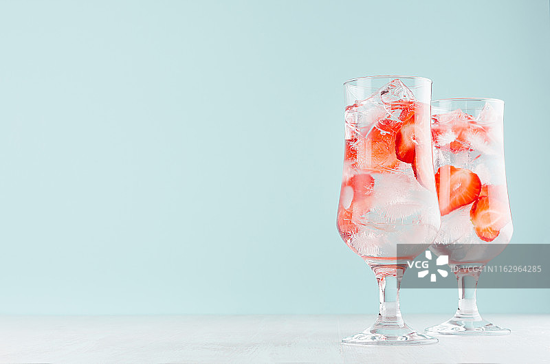 草莓柠檬水，熟透的浆果片，冰块和矿泉水在两个优雅的杯子在白色木桌上，薄荷色墙。图片素材