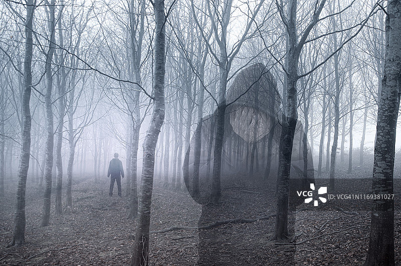 一个戴兜帽的人双手抱头的艺术双重曝光，站在一个阴森的，有雾的冬天的森林里。有个人站在后面。图片素材