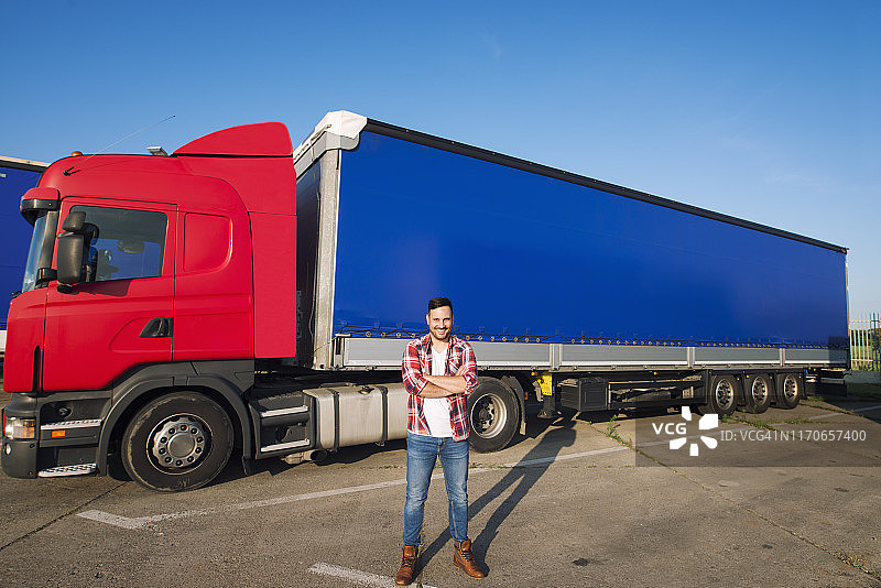 美国职业卡车司机的肖像，穿着休闲服装和靴子站在卡车前面与长拖车。运输服务。图片素材