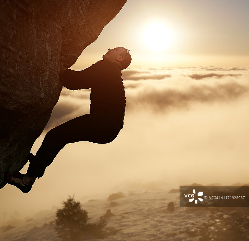 日落时，危险的男性站在陡峭的岩石上。坚持不懈的攀岩者向上看，准备实现他的目标图片素材
