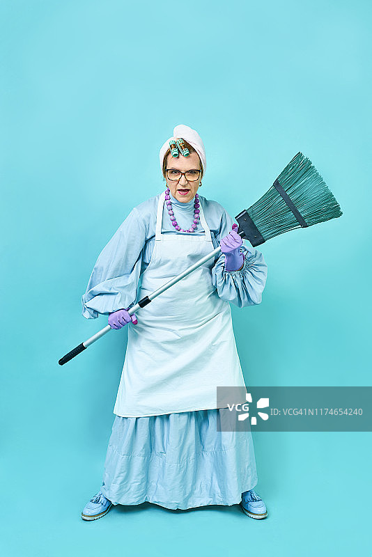 清洁女工的乐趣。滑稽的老家庭主妇拿着扫帚鬼混。全身隔离蓝图片素材
