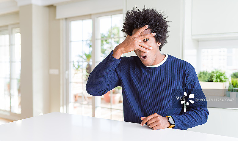 年轻的非洲裔美国男子穿着休闲毛衣坐在家里窥视在震惊覆盖的脸和眼睛用手，透过手指看与尴尬的表情。图片素材