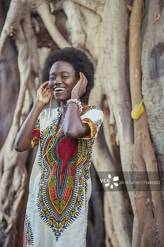 微笑的年轻女子站在树干前，戴着耳机图片素材