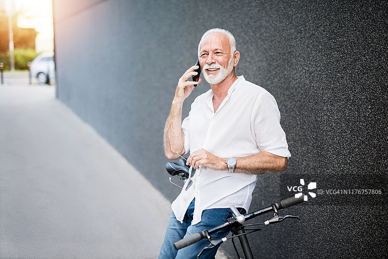 快乐健康优雅的老人靠在他的自行车上，在城市里打电话。图片素材