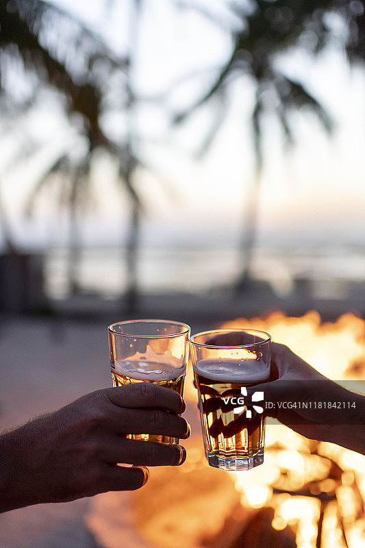 一对夫妇在热带海滩的篝火前欢呼啤酒在日落图片素材
