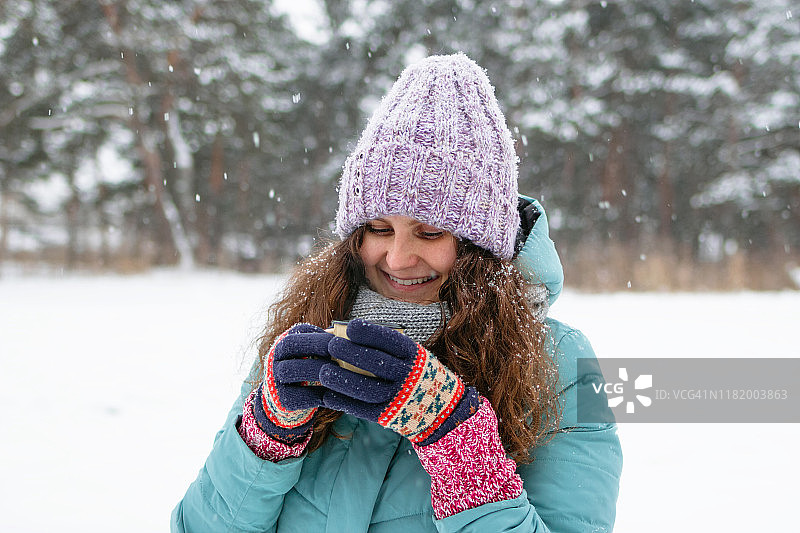美丽微笑的女人，长长的卷发，蓝色的冬季夹克和编织多彩的手套，手里拿着一杯热茶，在冬天的雪林散步。图片素材
