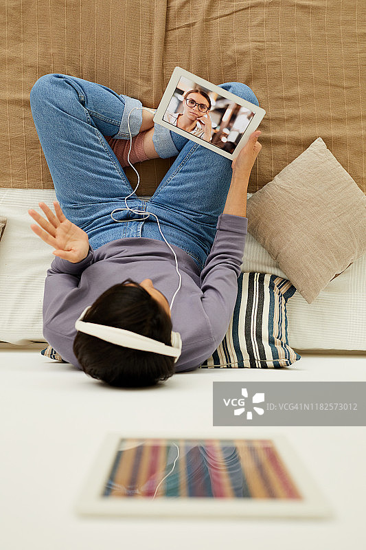 上图中，穿着牛仔裤的年轻女子坐在床上，打着手势与朋友在平板电脑上通过互联网应用聊天图片素材